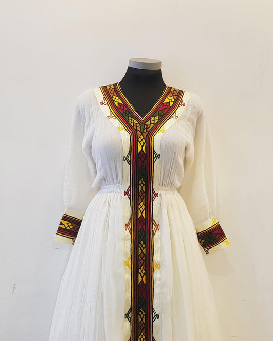 A white long-sleeve Ethiopian outfit displayed on a black mannequin torso against a plain off-white wall. The dress features a V-neckline and a full skirt, decorated with prominent vertical and horizontal geometric embroidery in red, yellow, and black along the neckline, center front, and cuffs.