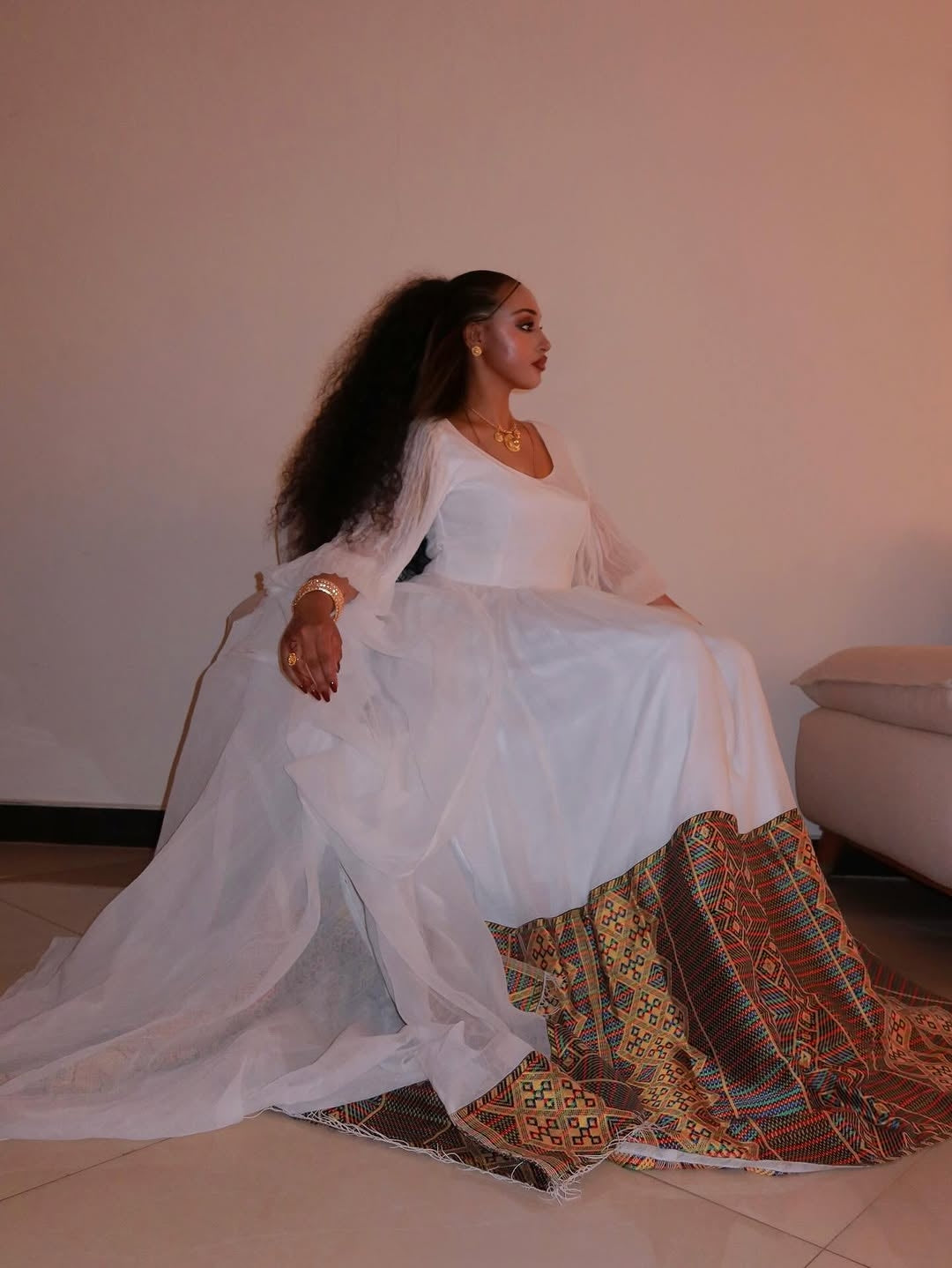 Woman in a white habesha kemis with a colorful patterned skirt sitting on a couch.
