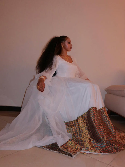 Woman in a white habesha kemis with a colorful patterned skirt sitting on a couch.