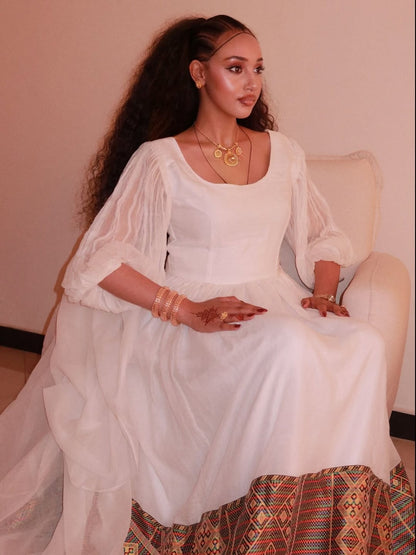 Woman in a white habesha kemis with long sleeves sitting on a chair.