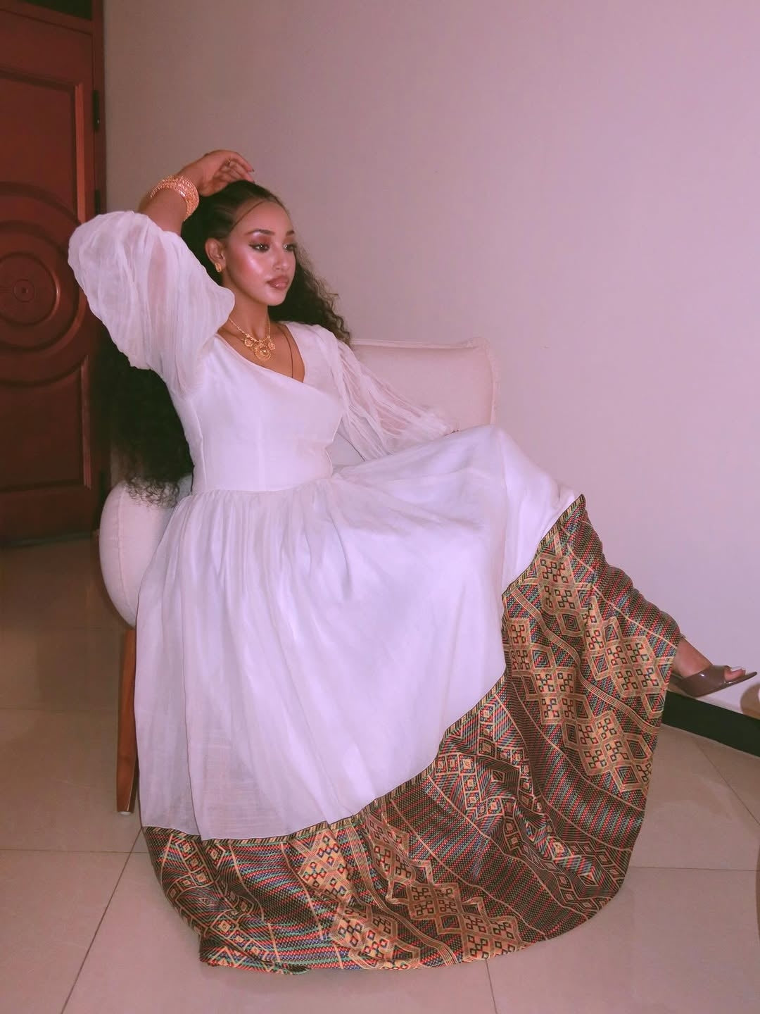 Woman in a white habesha kemis with a patterned skirt sitting on a chair.