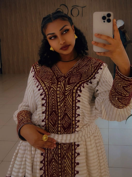 A woman takes a mirror selfie wearing a cream-colored long-sleeve Ethiopian traditional clothing with intricate dark brown and beige geometric embroidery. She is holding a white smartphone to capture the outfit, set against an indoor background with a tiled floor and a wooden-paneled wall.