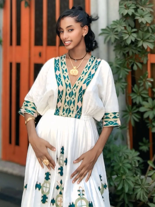 Woman wearing a white habesha dress with green and beige patterns, standing outdoors.