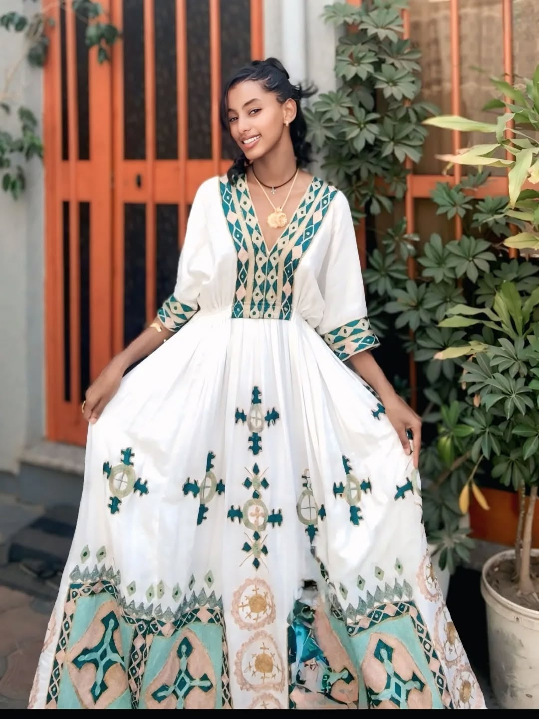 Woman wearing a white habesha dress with green and blue patterns standing outdoors.