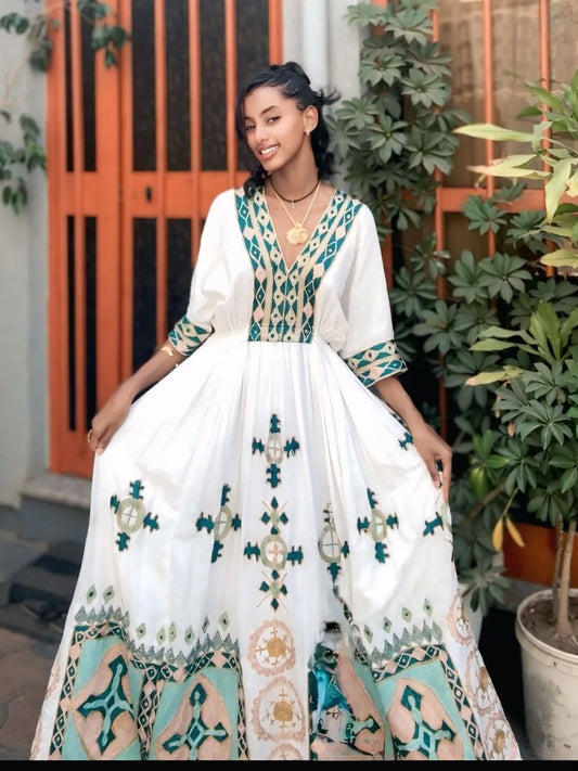Woman wearing a white habesha dress with green and blue patterns standing outdoors.