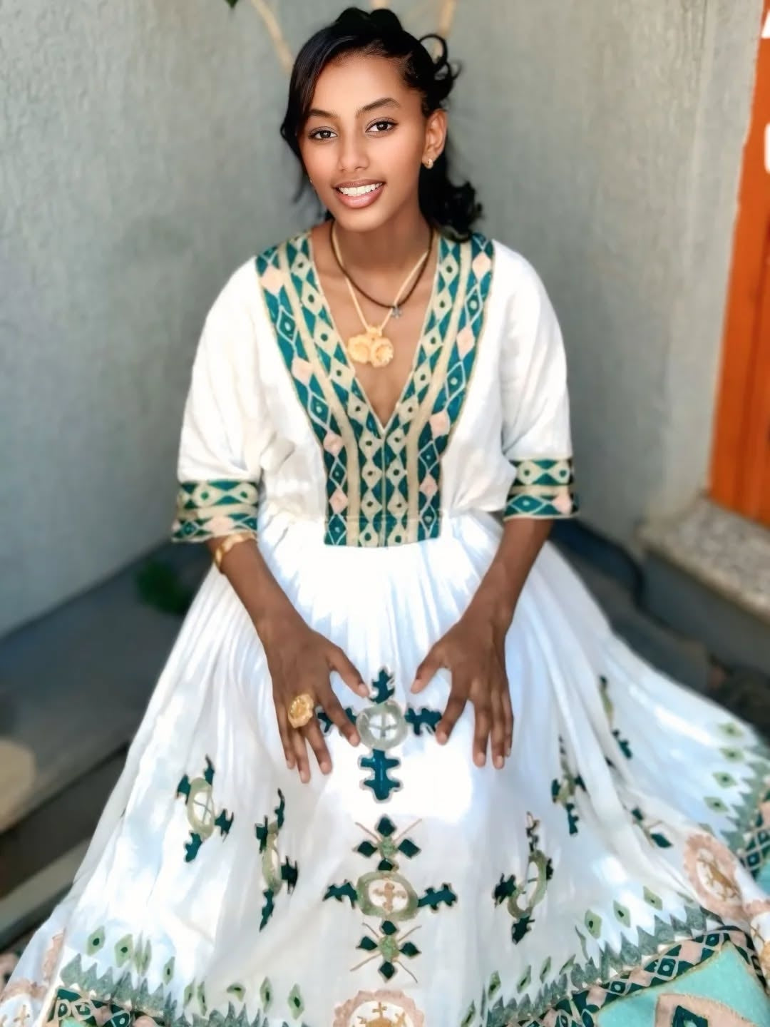 Woman wearing a white habesha dress with green and blue patterns, sitting against a light-colored wall.