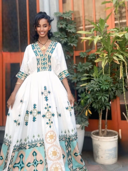 Woman in a white habesha dress with blue and green patterns standing outdoors with plants in the background
