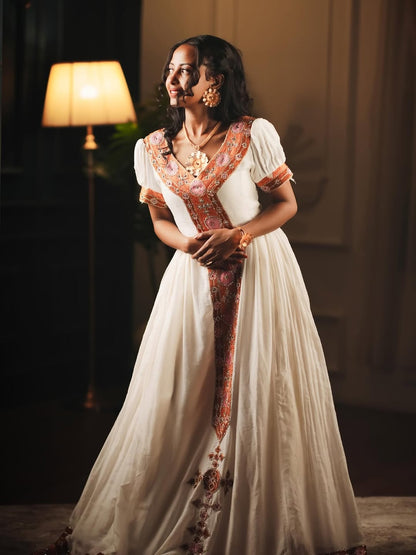 Woman in a white and orange traditional Habesha dress  standing in a dimly lit room.