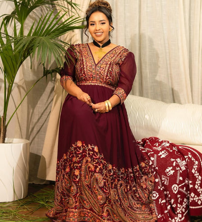 Woman in a burgundy Habesha Chiffon with intricate patterns standing indoors next to a white couch and plant.