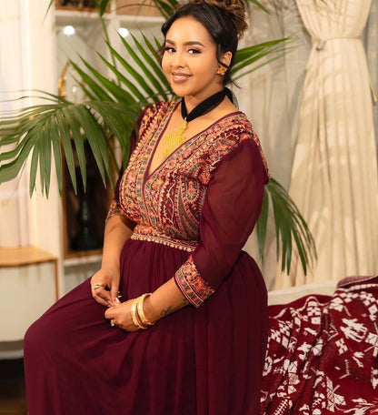 Woman wearing a maroon traditional Habesha Chiffon with intricate patterns, sitting indoors.