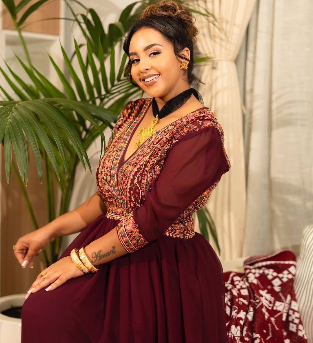 Woman in a burgundy traditional Habesha Chiffon with intricate patterns, sitting indoors.