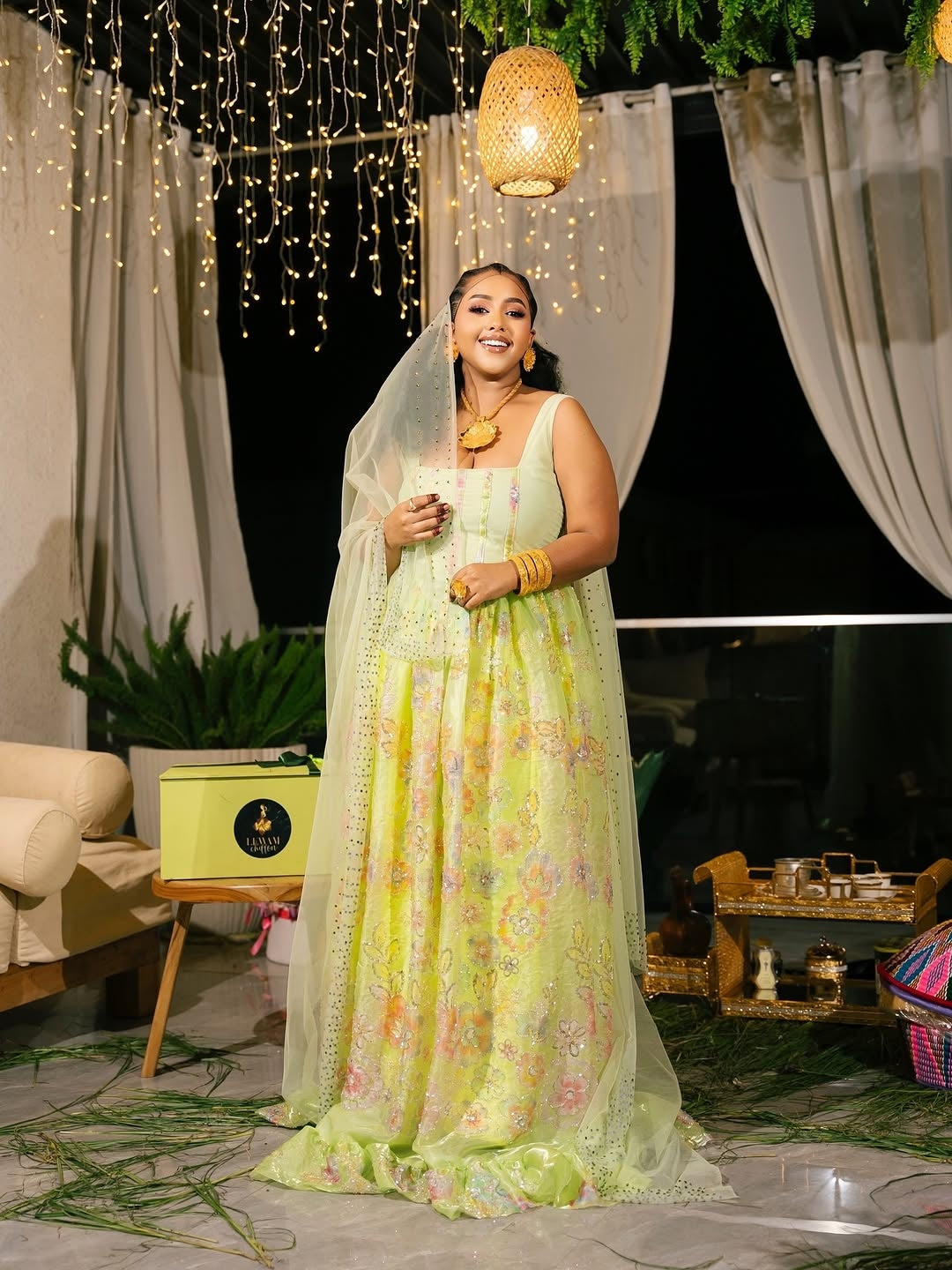 Woman in a light green traditional Habesha Chiffon standing in an outdoor setting with string lights and plants.