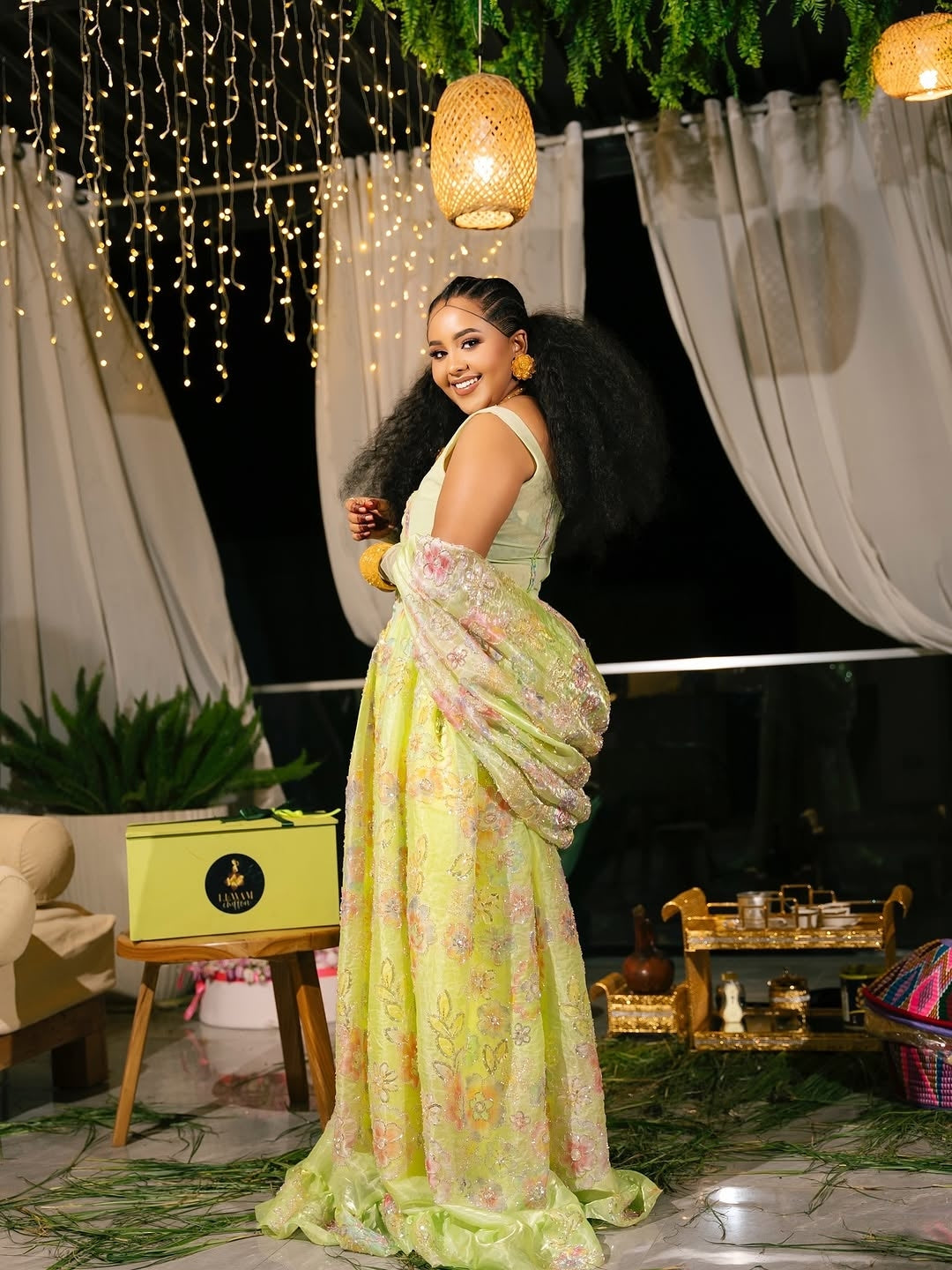 Woman in a floral Habesha Chiffon standing in an outdoor setting with string lights and decor.