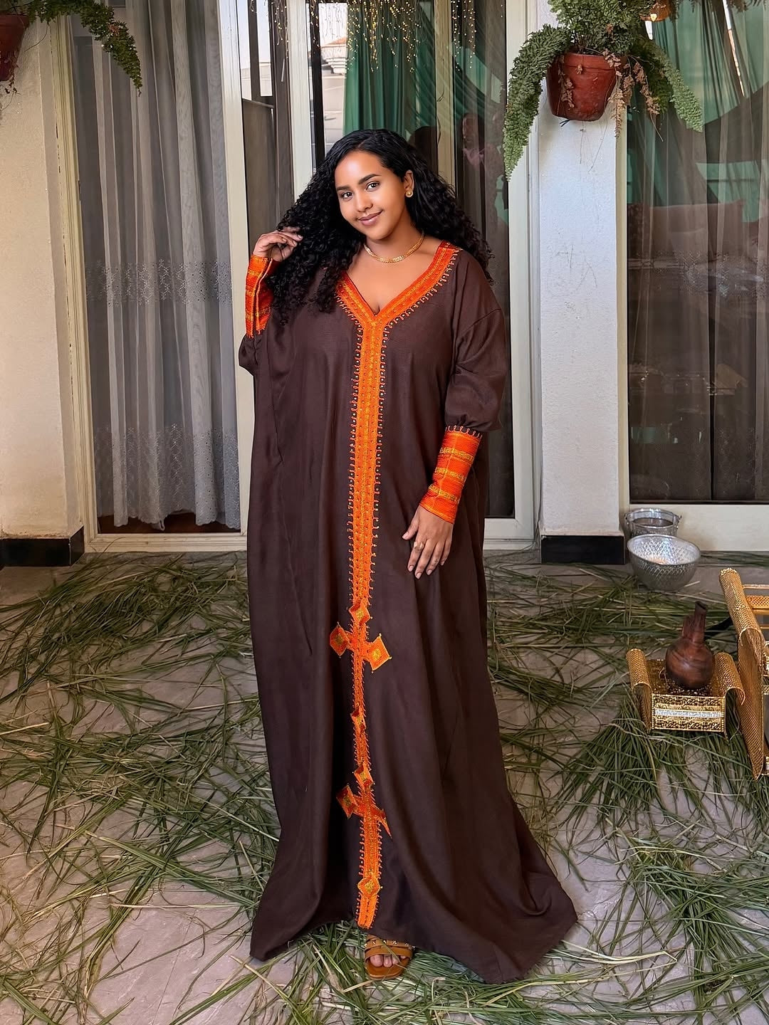 Woman wearing a brown Habesha Dress with orange accents standing in a room with decorative elements.