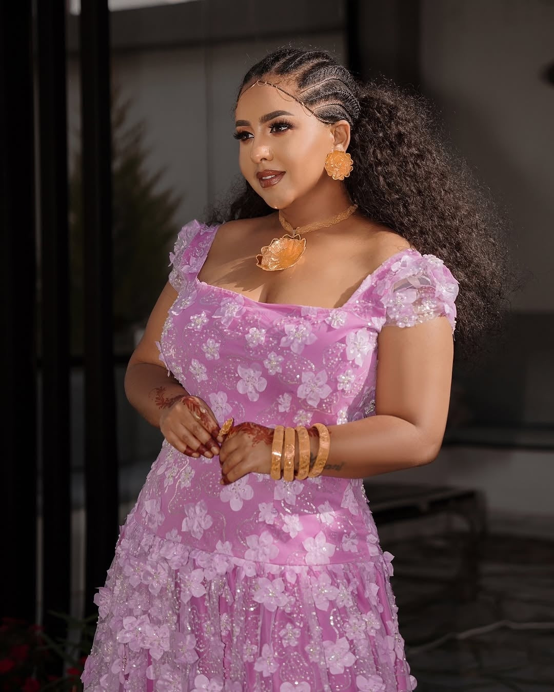Woman in a pink floral Habesha Shiffon with jewelry, standing against a blurred background