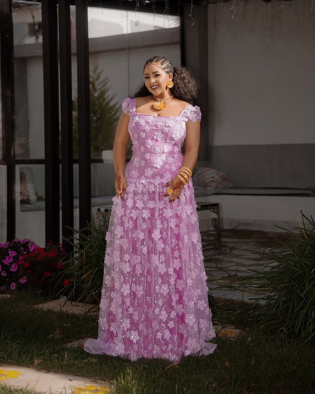 Woman in a pink floral Habesha Shiffon standing outdoors with plants and a building in the background