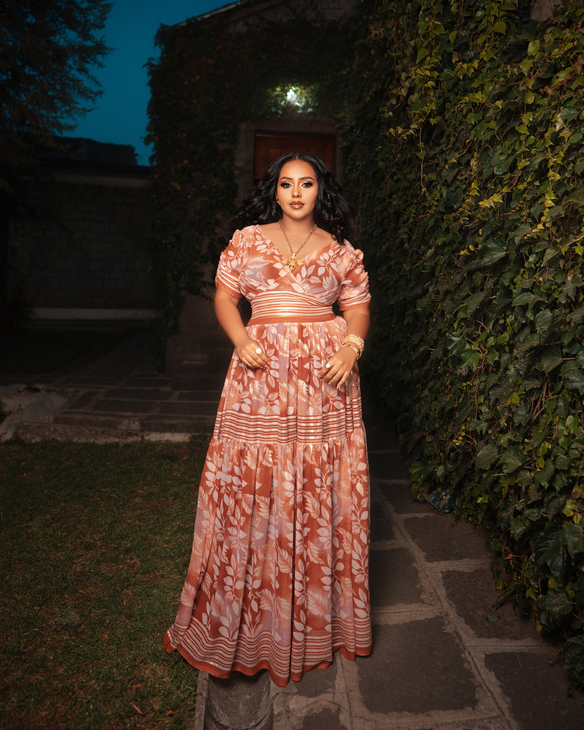 A woman stands outdoors on a stone path wearing a long, flowing Ethiopian Dress Chiffon with a terracotta and cream floral pattern. The dress has short puff sleeves, a V-neckline, and a fitted waist with a matching belt.