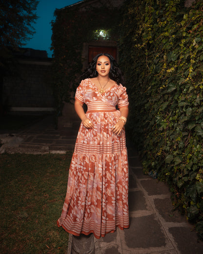 A woman stands outdoors on a stone path wearing a long, flowing Ethiopian Dress Chiffon with a terracotta and cream floral pattern. The dress has short puff sleeves, a V-neckline, and a fitted waist with a matching belt.