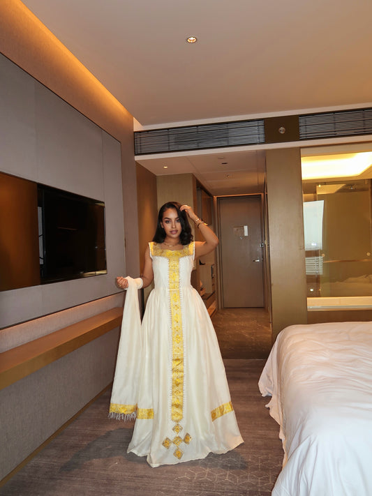 A woman modeling a white traditional Ethiopian national dress with gold embroidery in a modern hotel room.