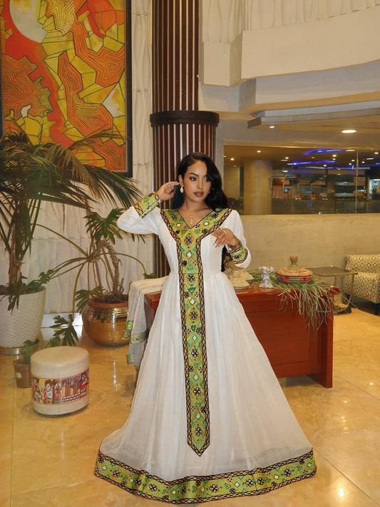 Woman in a traditional white and green dress with intricate patterns in a decorated indoor setting.