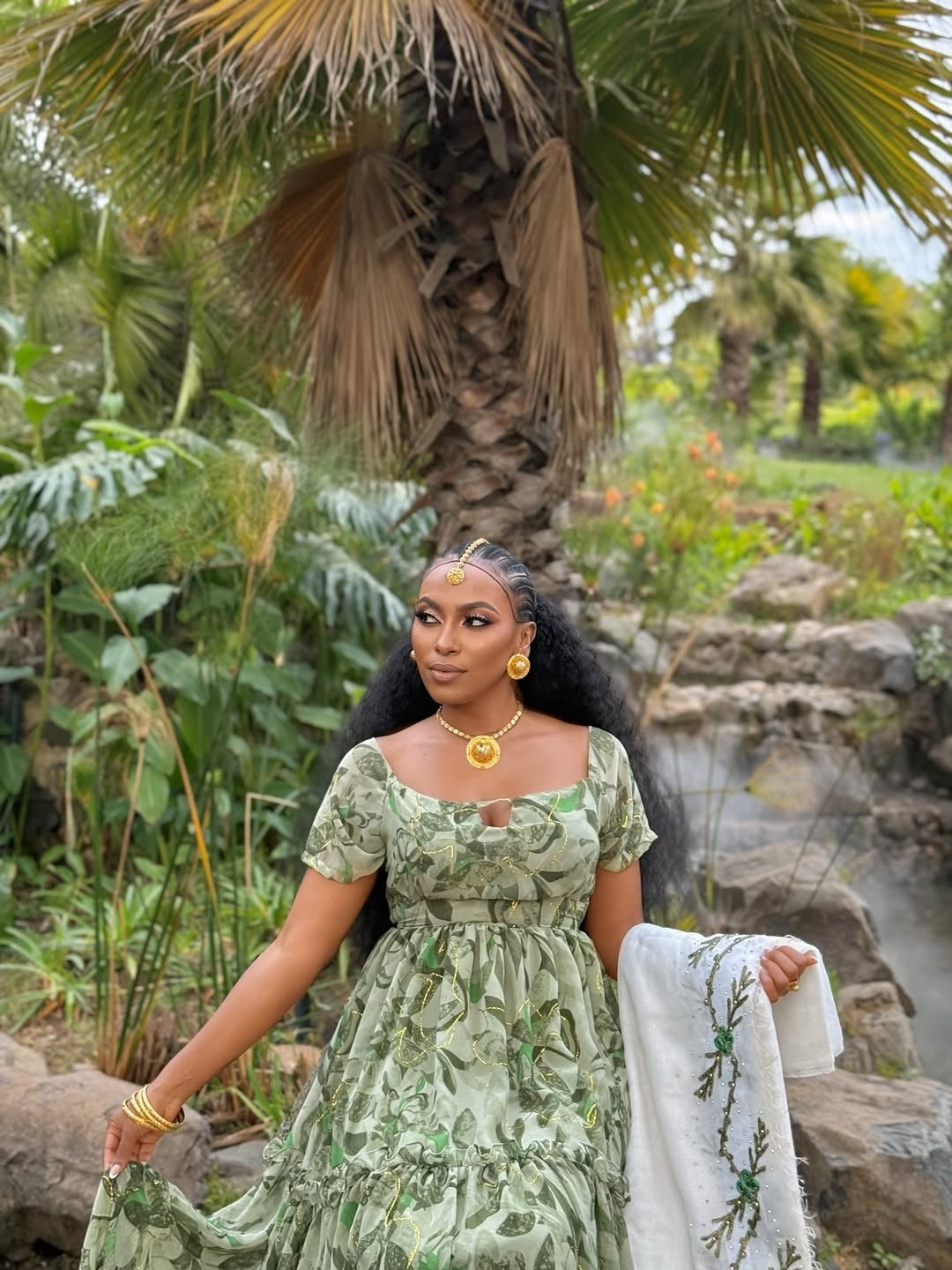 Woman in a green dress standing in a garden with palm trees and rocks.