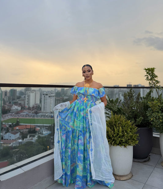 A woman stands on a rooftop terrace wearing a long off-the-shoulder maxi chiffion Habesha  with a blue, green, and yellow tie-dye pattern, holding a sheer white dupatta. The background features a cityscape at dusk with buildings, a running track, and potted plants.