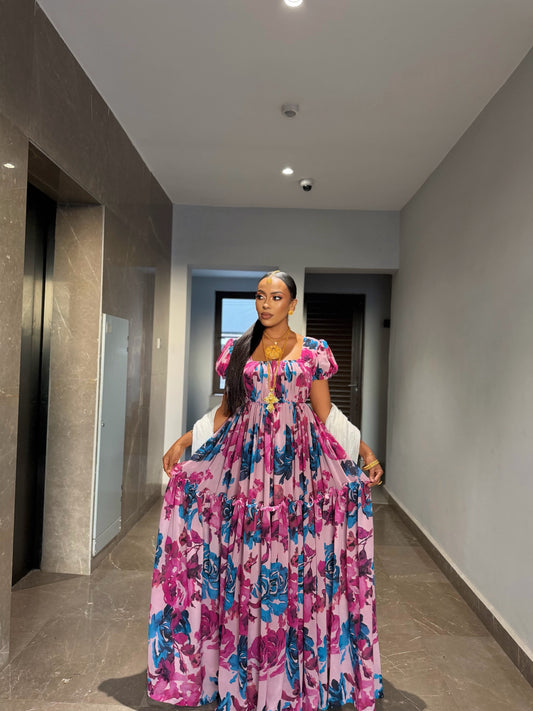 A woman standing in a hallway wearing a pink habesha chiffon dress with a vibrant blue and purple floral pattern, styled with traditional gold jewelry 