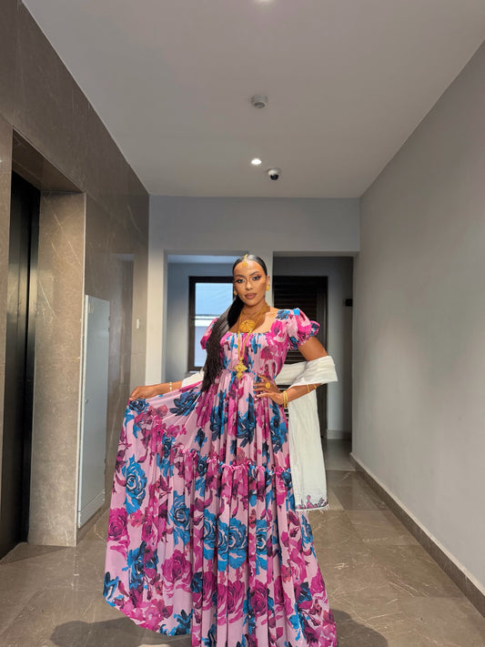 A woman standing in a hallway wearing a pink habesha chiffon dress with a vibrant blue and purple floral pattern, styled with traditional gold jewelry 