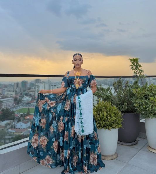 A woman stands on a rooftop terrace at sunset wearing a floor-length teal floral habehsa chiffion with off-the-shoulder sleeves and holding a white embroidered sash. The background shows a city skyline, potted plants, and a cloudy sky.