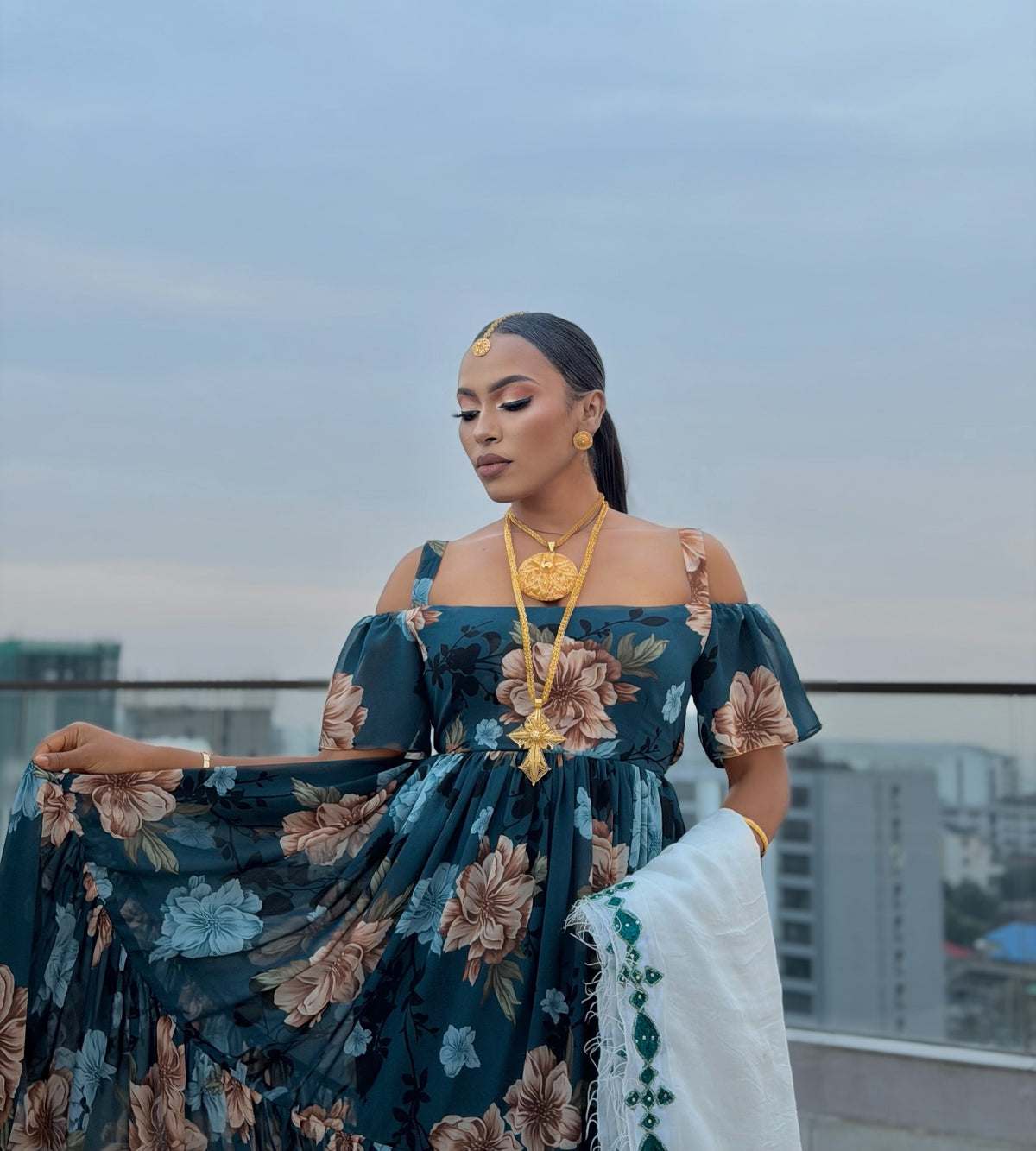 A woman stands on a rooftop wearing a dark teal off-the-shoulder floral habesha chiffion with large beige and light blue flowers, gold jewelry, and a white draped fabric. City buildings and a pale sky are visible in the background.