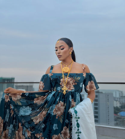 A woman stands on a rooftop wearing a dark teal off-the-shoulder floral habesha chiffion with large beige and light blue flowers, gold jewelry, and a white draped fabric. City buildings and a pale sky are visible in the background.