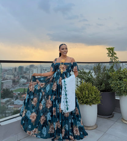 A woman stands on a rooftop terrace wearing a voluminous off-the-shoulder teal floral habesha chiffionwith a white embroidered sash, holding out the skirt to show its scale. The background features a city skyline at sunset, potted green plants, and a glass railing.