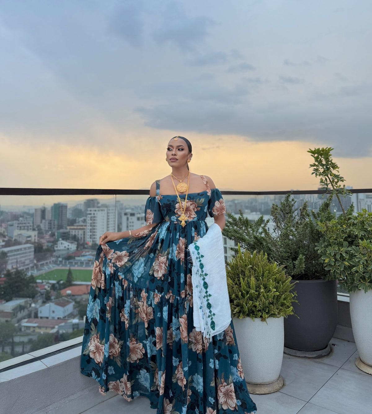 A woman stands on a rooftop terrace wearing a floor-length off-the-shoulder teal blue floral habesha chiffionwith a white embroidered shawl, holding out the skirt to show its volume. The background features a city skyline at dusk with potted plants.
