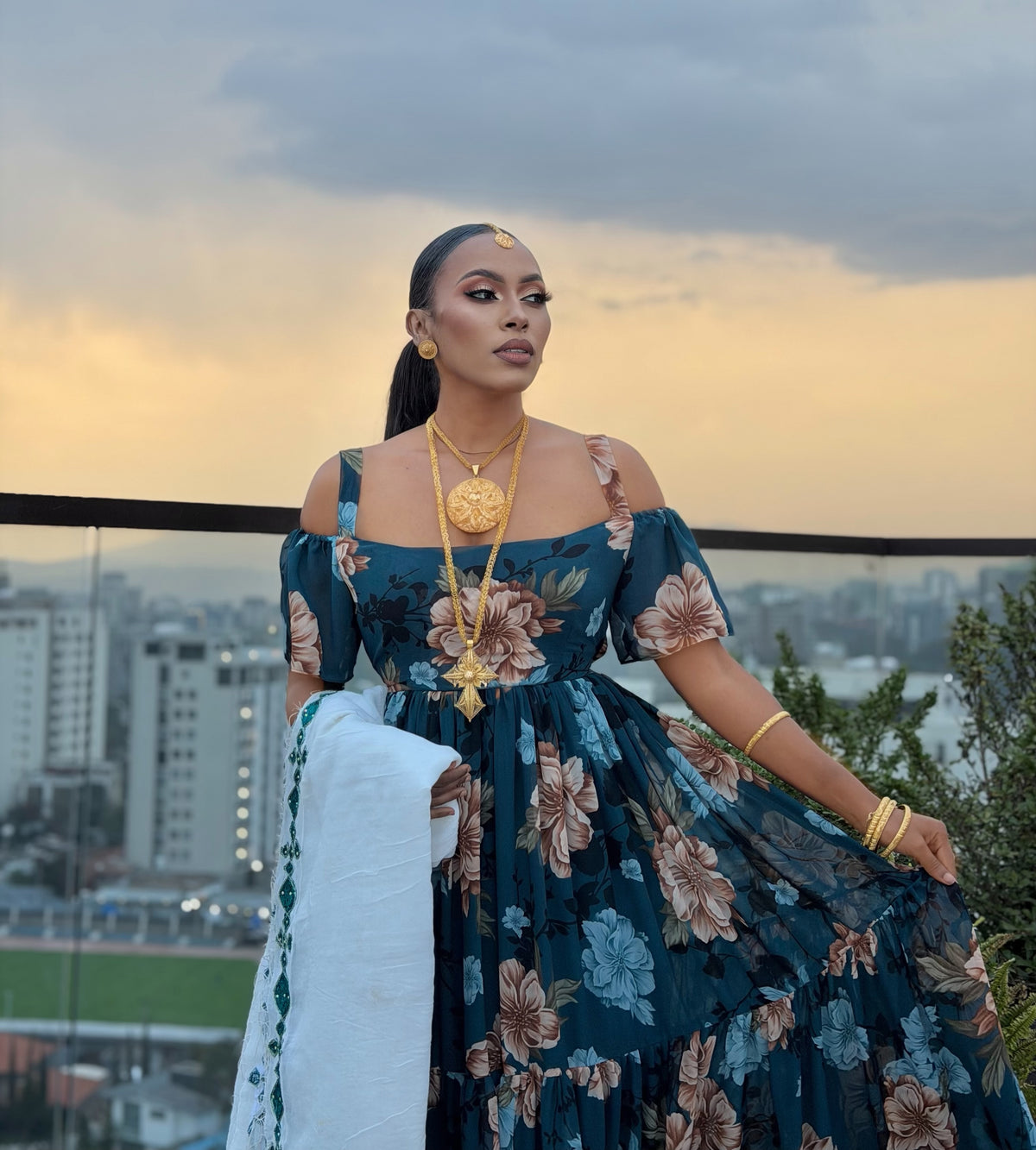 A woman stands outdoors on a balcony at sunset wearing a dark teal floral off-the-shoulder habesha chiffion with a full skirt, layered gold jewelry, and a white draped fabric. The background shows a city skyline and cloudy sky.