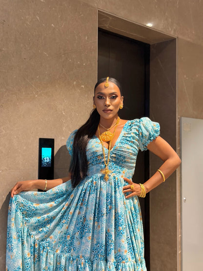 A woman wearing a light blue floral print maxi Habesha chiffion  with puff sleeves and a deep V-neckline, accessorized with gold jewelry, standing in front of a beige marble wall and elevator door.