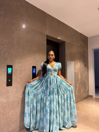 A woman wearing a light blue floral print ball Habesha chiffion  with gold trim, holding out the sides of the skirt to show its full volume, standing in a modern lobby with brown marble walls.