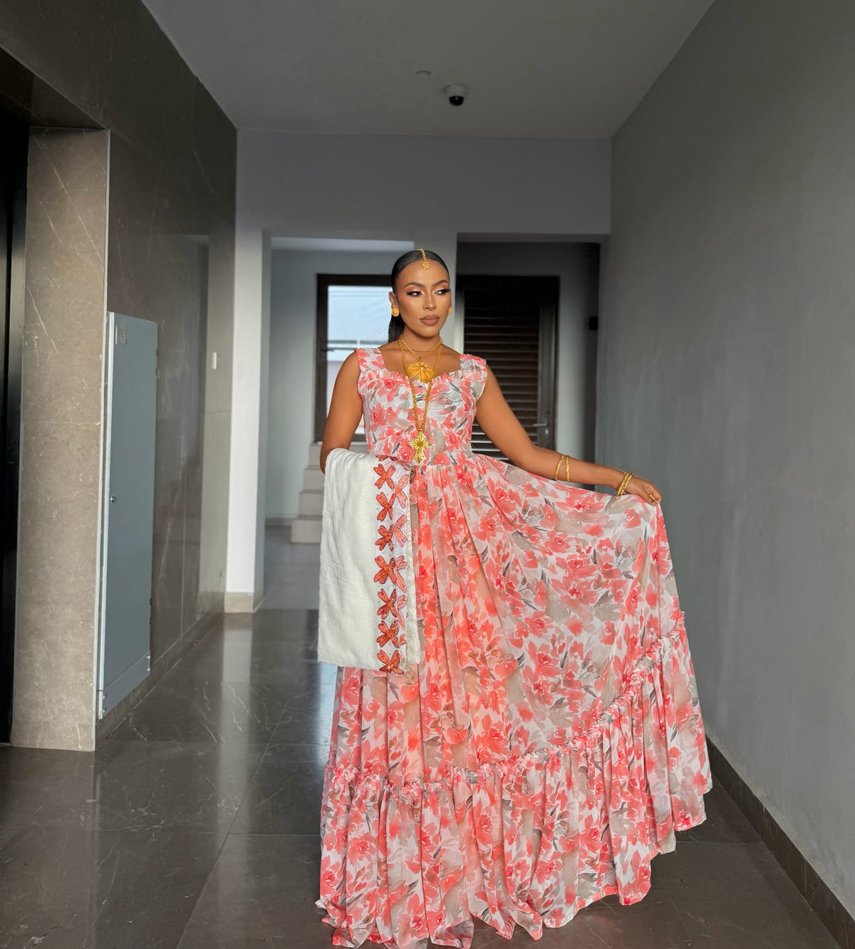 A woman stands in a hallway wearing a sleeveless, tiered Habesha chiffion with a pink and coral floral print on a white base, holding out one side of the skirt to show the drape and a white dupatta draped over her arm.