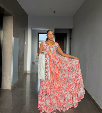 A woman stands in a hallway wearing a sleeveless, tiered Habesha chiffion with a pink and coral floral print on a white base, holding out one side of the skirt to show the drape and a white dupatta draped over her arm.