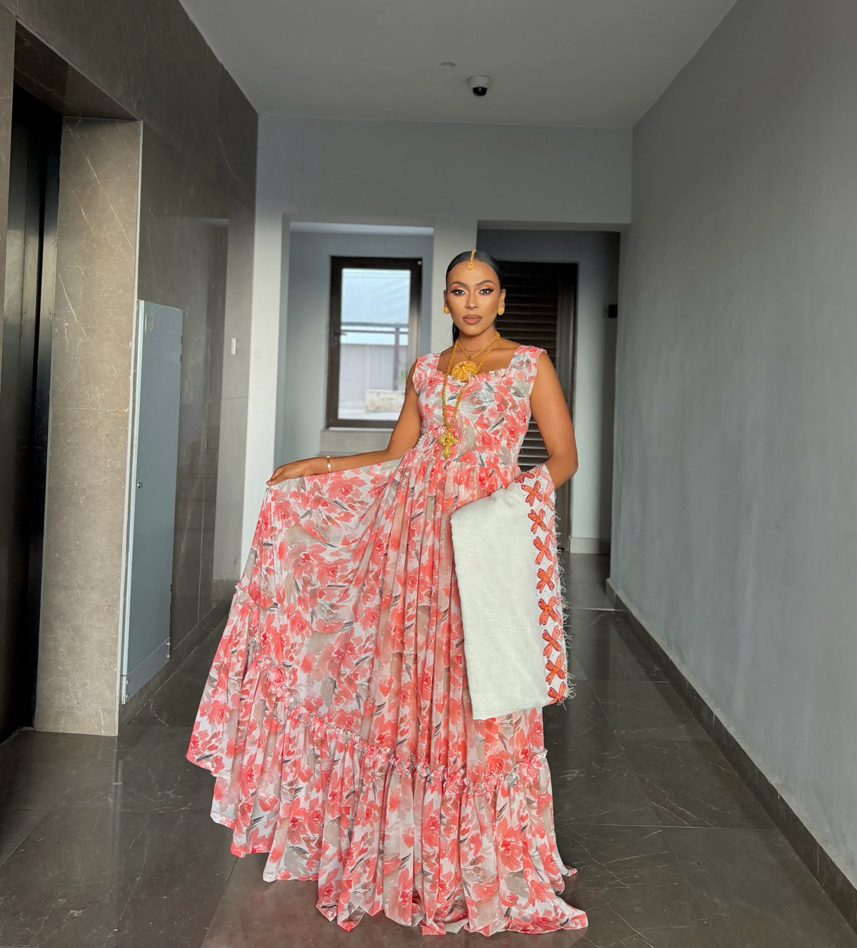 A woman stands in a hallway wearing a sleeveless, tiered maxi habesha chiffion  with a pink and coral floral print on a white base, holding a white dupatta.