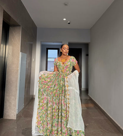 A woman stands in a hallway wearing a long floral printed Habesha chiffion  with a white dupatta draped over her arms. The dress has a green base with pink, yellow, and blue floral patterns, and she is wearing a gold necklace.