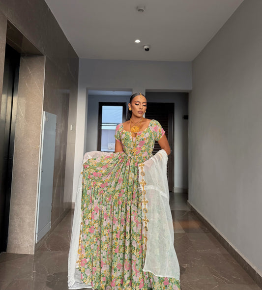 A woman stands in a hallway wearing a long floral printed Habesha chiffion  with a white dupatta draped over her arms. The dress has a green base with pink, yellow, and blue floral patterns, and she is wearing a gold necklace.