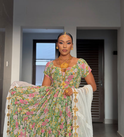 A woman stands indoors wearing a floral printed traditional Habesha chiffion  with a white dupatta featuring gold embroidered borders. She is also wearing gold jewelry, including a large pendant necklace, earrings, and a forehead ornament.