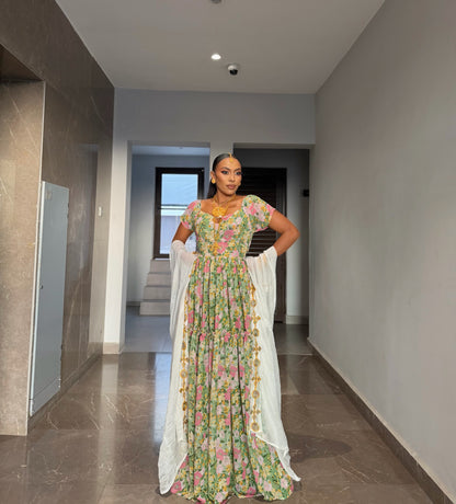 A woman stands in a hallway wearing a long floral printed Habesha chiffion  with a white draped scarf. The dress has a green base with pink, yellow, and blue floral patterns, and the background shows a modern interior with tiled floors and neutral walls.