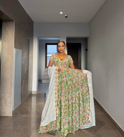 A woman stands in a hallway wearing a long floral Habesha chiffion  with a white dupatta. The dress has a green base with pink and yellow floral patterns and gold embellishments.