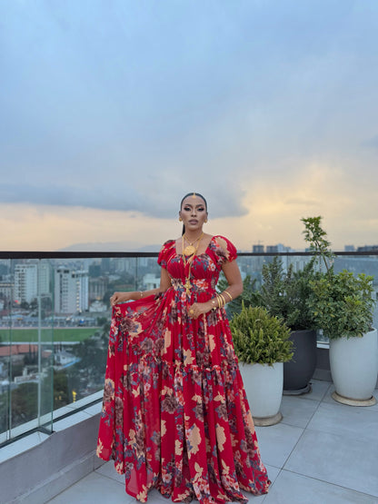 Woman in a red, tiered floral chiffon Habesha dress on a balcony with a city skyline background.