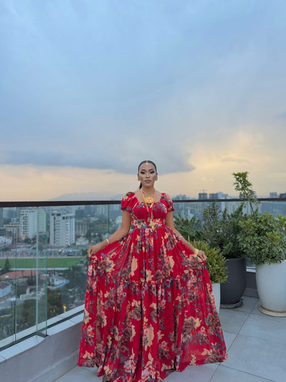 Woman in a red, tiered floral chiffon Habesha dress on a balcony with a city skyline background.