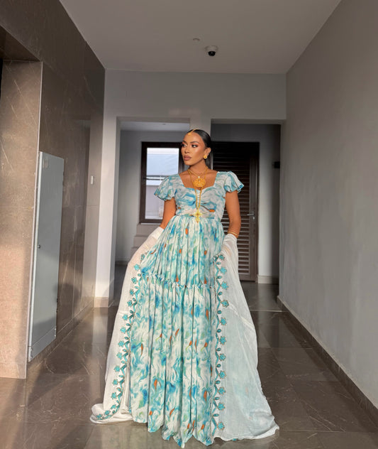 A woman wearing a light blue and white floral print traditional habesha chiffon with a gold waist tie, standing in a hallway.