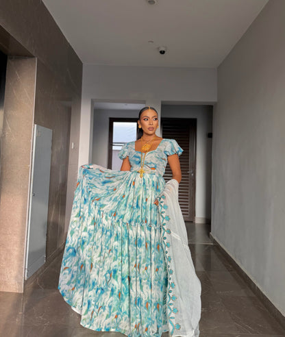A woman stands in a hallway wearing a traditional habesha chiffon  with a blue, white, and gold abstract print, holding the skirt out to show its volume. The dress features a matching light blue dupatta and a gold necklace.