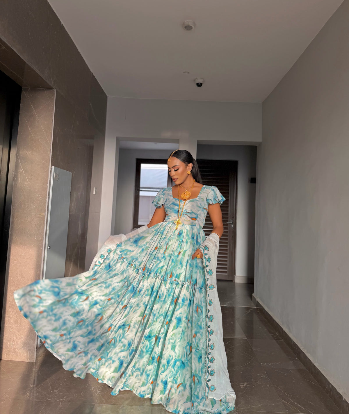 A woman wearing a long, flowing blue and white patterned habesha chiffon  with puff sleeves, standing in a hallway and holding out the skirt to show its volume.