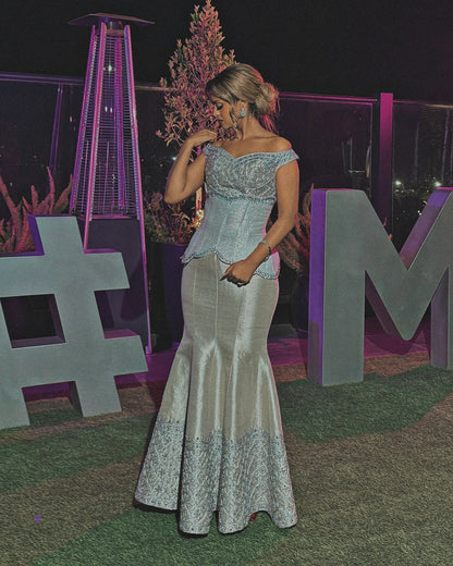 A woman wearing a formal off-the-shoulder evening Traditional Ethiopian Clothing with a light blue lace bodice and a shimmering silver-gray satin mermaid skirt, standing at night in front of decorative letters and a purple-lit heater.